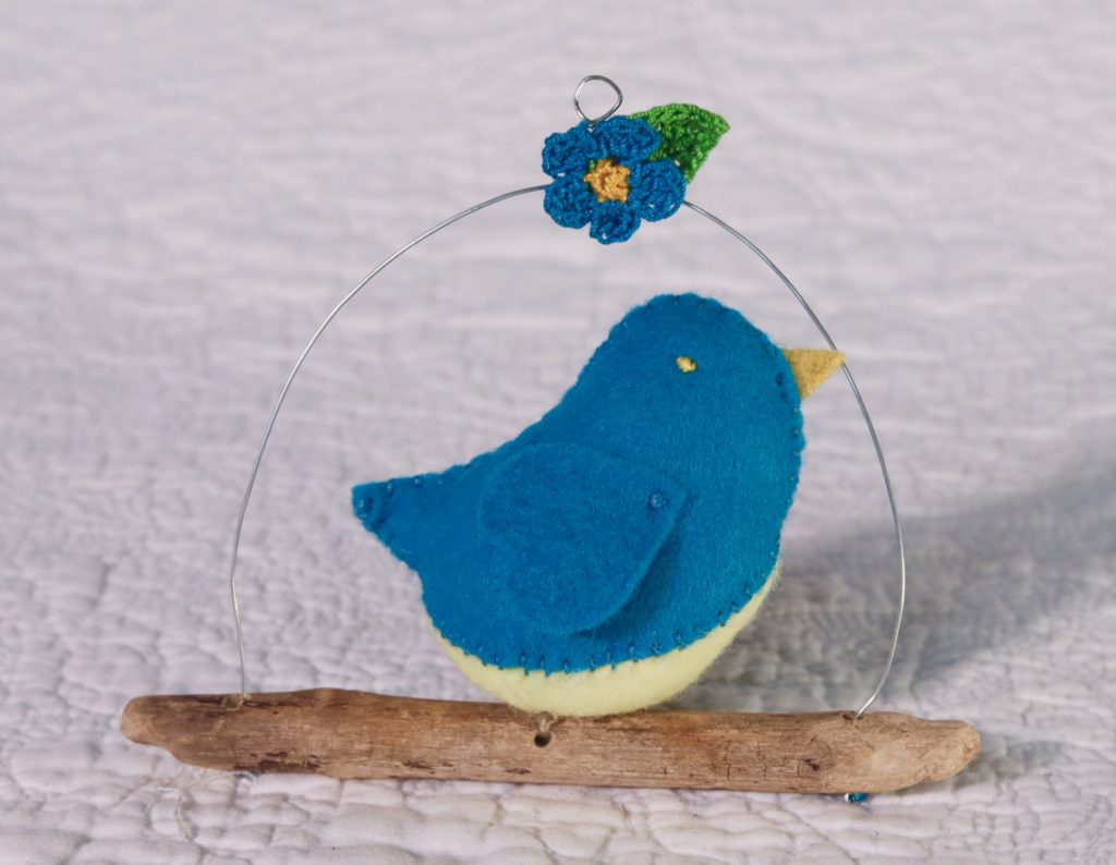 Single hand sewn felt bird sat on a driftwood branch with wire hanging hoop and small crocheted detail.